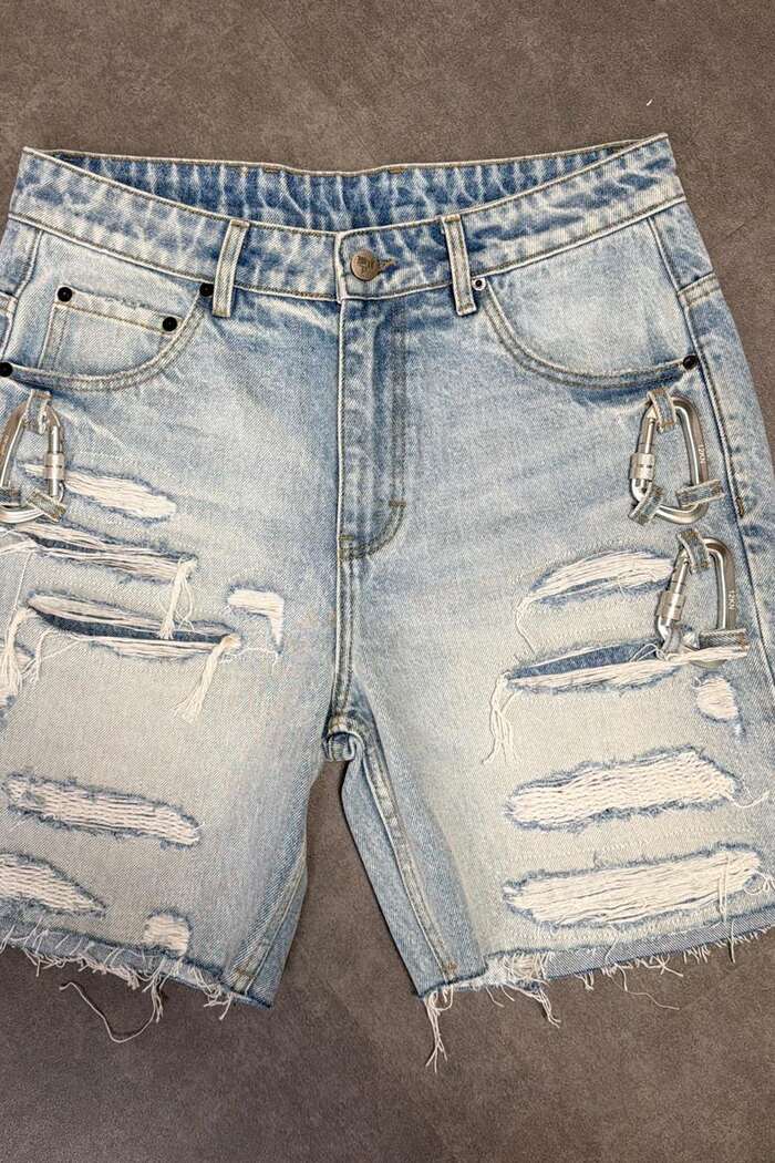 A pair of distressed light-wash denim shorts lying flat on a concrete floor, frayed at the hem, with silver carabiner hardware clipped through both hip pockets.